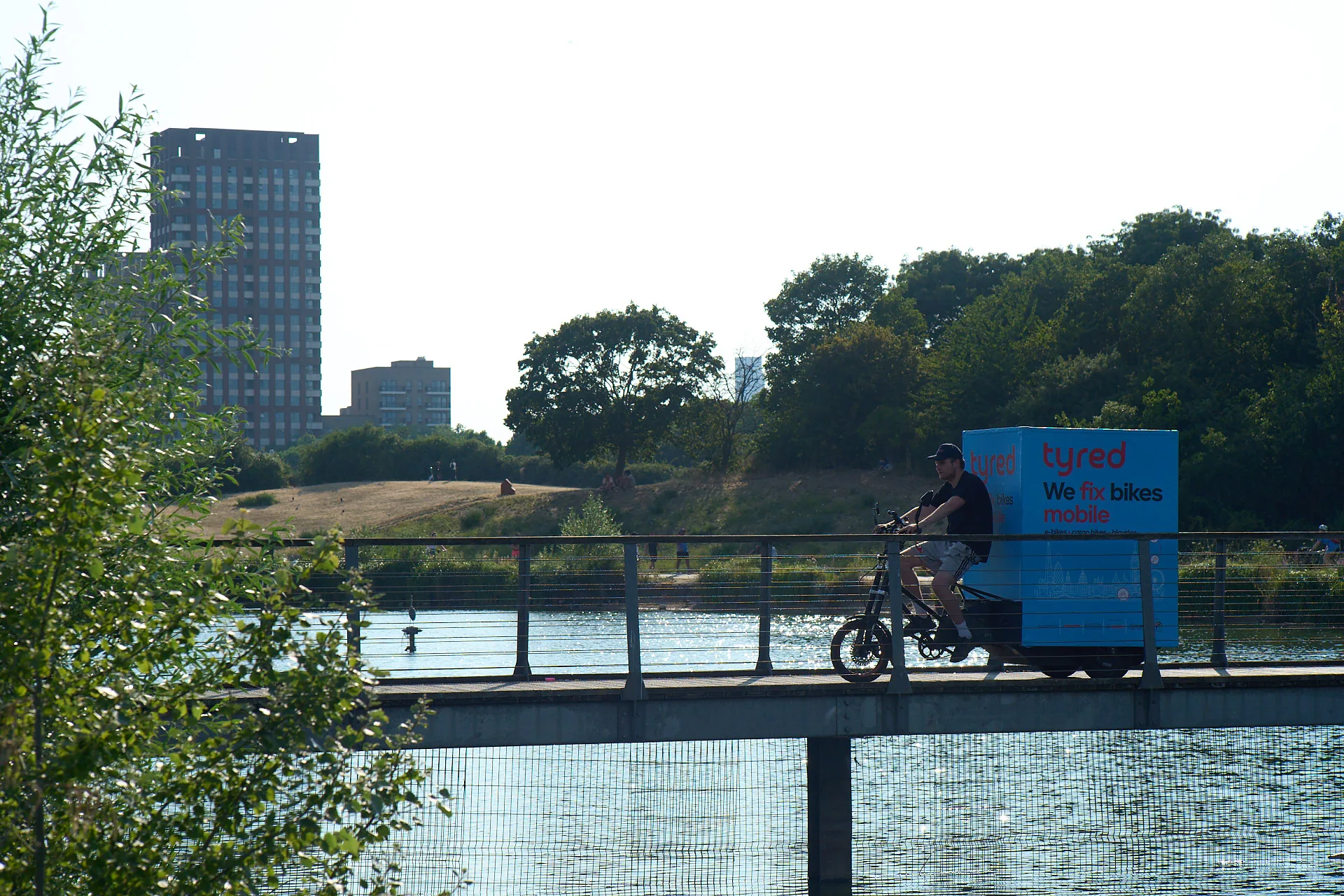 A Tyred mechanic riding a branded cargo bike along a London greenway.